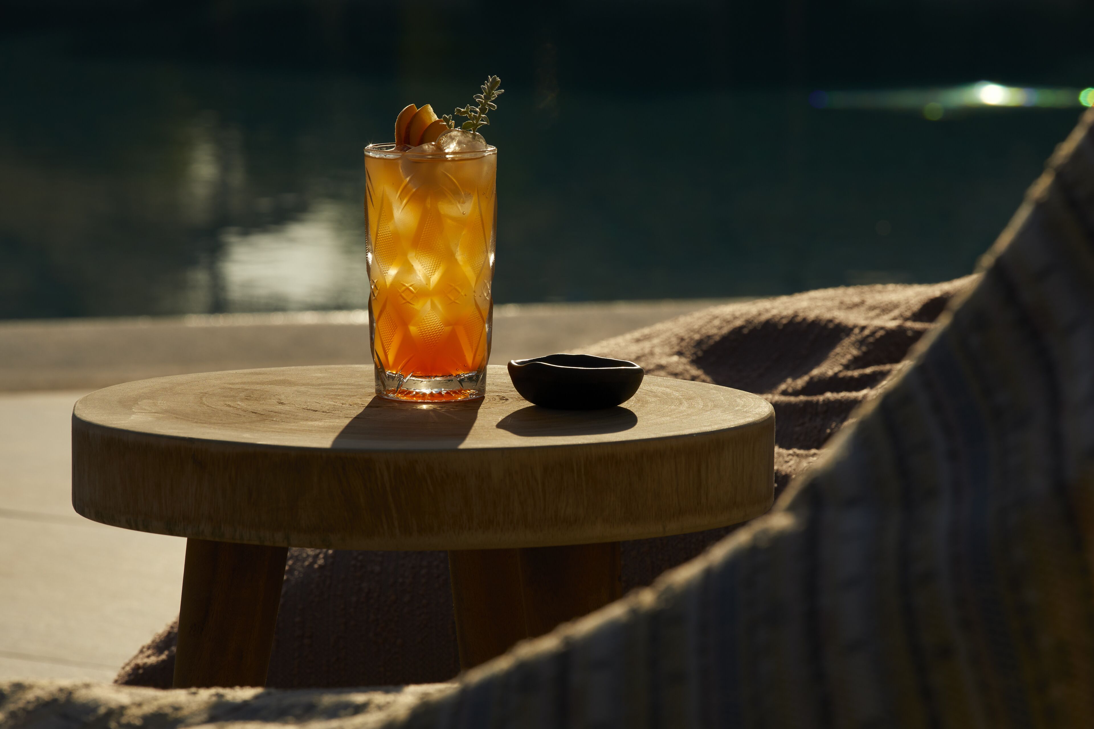 Un vaso de bebida color ámbar sobre una mesa de madera junto a una piscina, con un tazón pequeño a un lado.