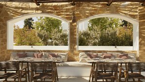 Un área de estar al aire libre con paredes de piedra, muebles de madera y grandes ventanales con vistas a un jardín.