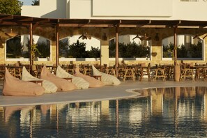 Un área junto a la piscina con varios espacios de descanso, incluyendo sillones tipo orejero y sillas, bajo un porche cubierto con paredes de piedra y amplios ventanales.