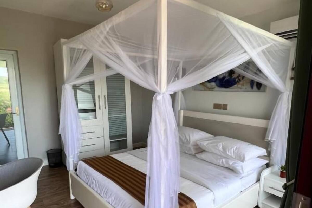 A white four-poster bed with a canopy, a wooden floor, a white chair, and a small bedside table with a plant.