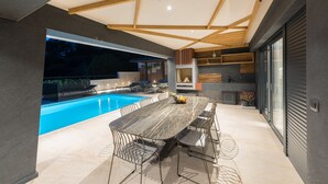 Un espace de restauration extérieur moderne, doté d'une piscine, de poutres en bois au plafond et d'une grande table entourée de chaises.