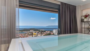 Une chambre d’hôtel moderne dotée d’une grande fenêtre donnant sur la ville, d’une piscine entourée de parois en verre et d’un décor minimaliste.