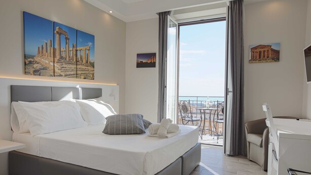 A modern hotel room with a large bed, a desk, and a balcony with a view of the city. - Esseneto Rooms (Agrigento)