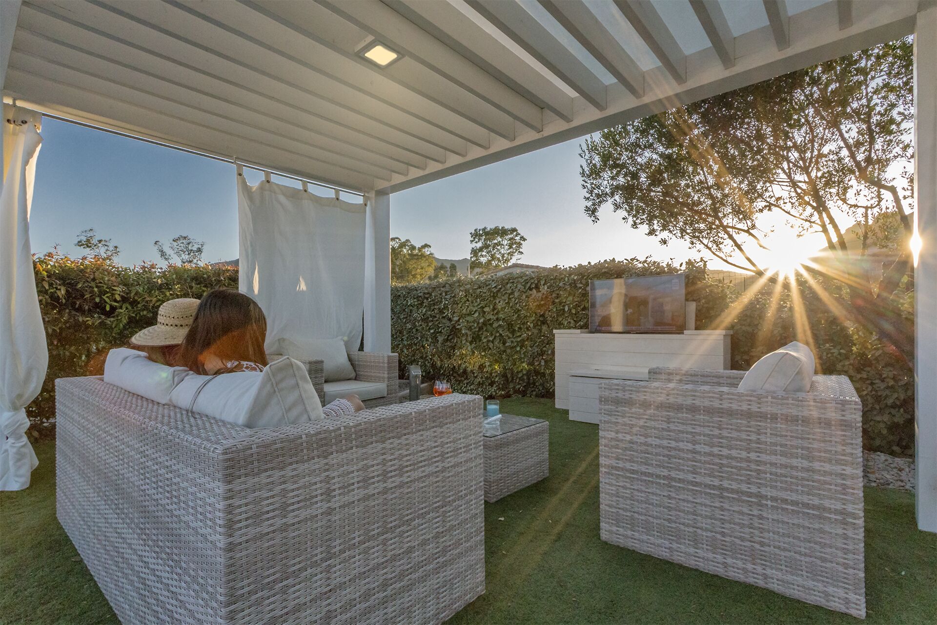 A covered outdoor seating area with wicker furniture, a glass table, and a TV.
