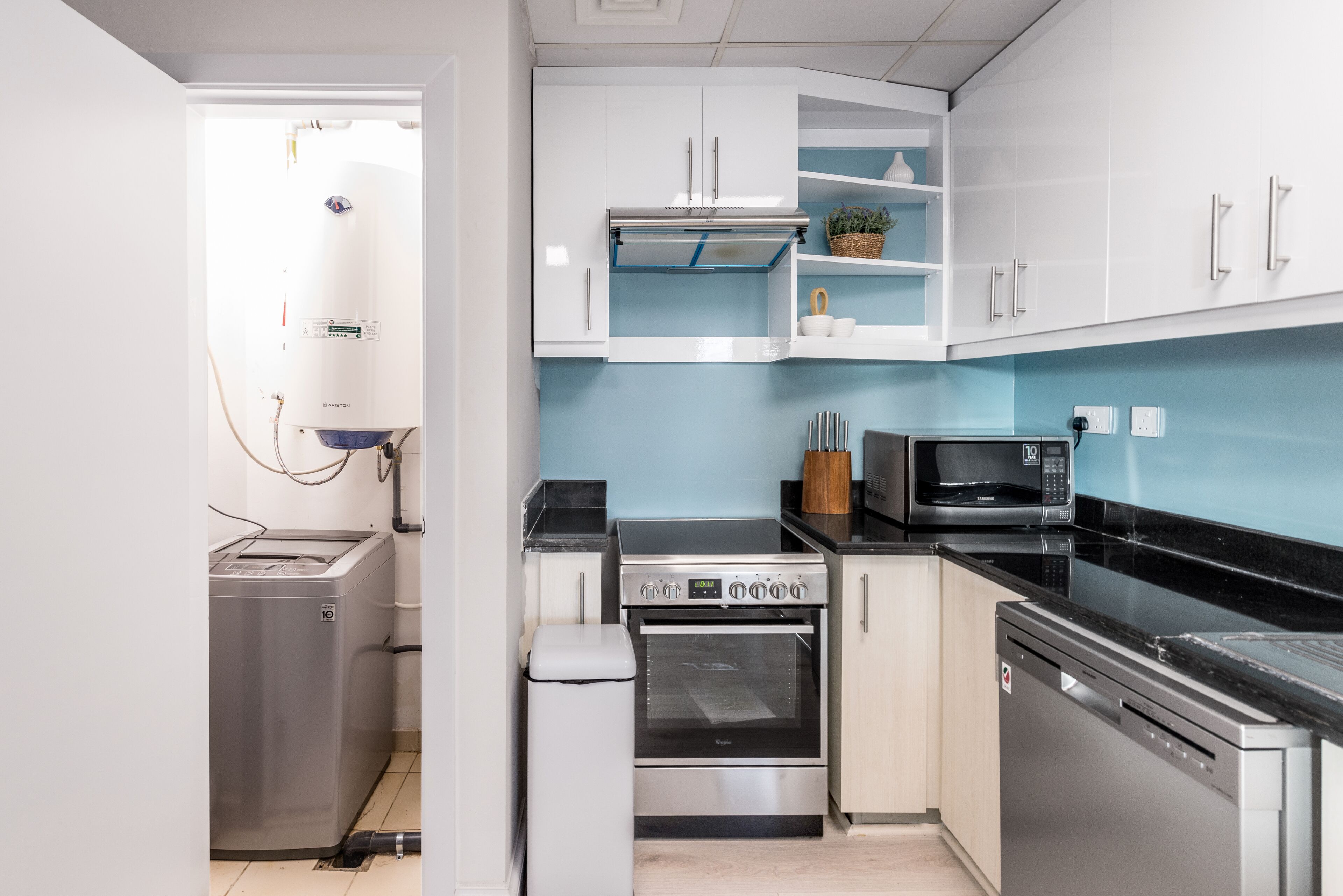 Una cocina compacta con armarios blancos, microondas, lavavajillas y calentador de agua.