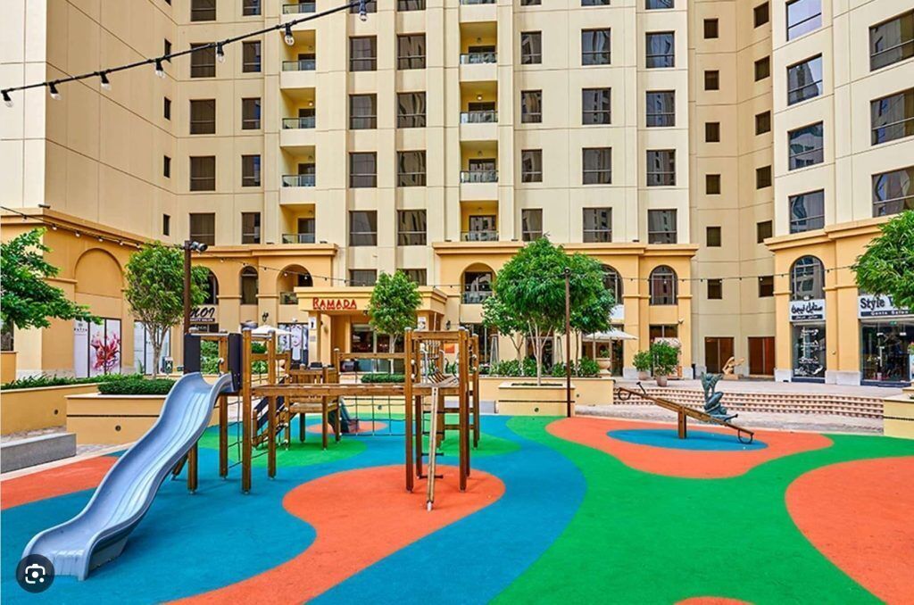 Un parque infantil cubierto con zonas de juego coloridas y un tobogán, rodeado por un edificio de varios pisos y balcones.