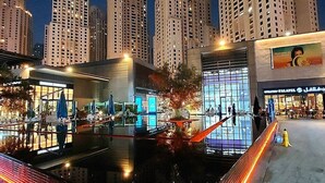 Un paisaje urbano nocturno con edificios altos, una plaza bien iluminada con un estanque reflectante y un edificio moderno con fachada de vidrio.