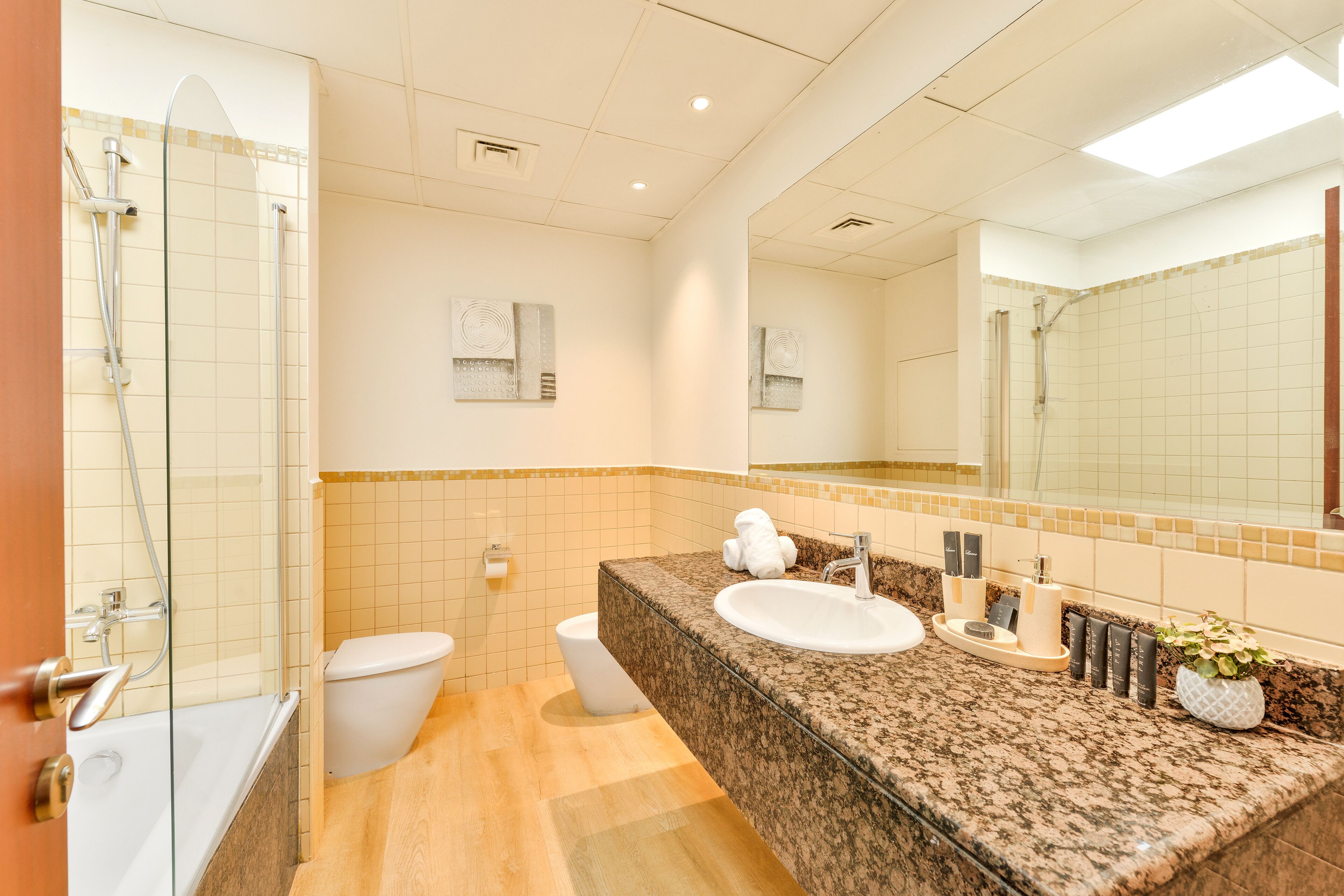Un baño moderno con ducha envuelta en vidrio, inodoro blanco, lavamanos con encimera de granito y un espejo grande.