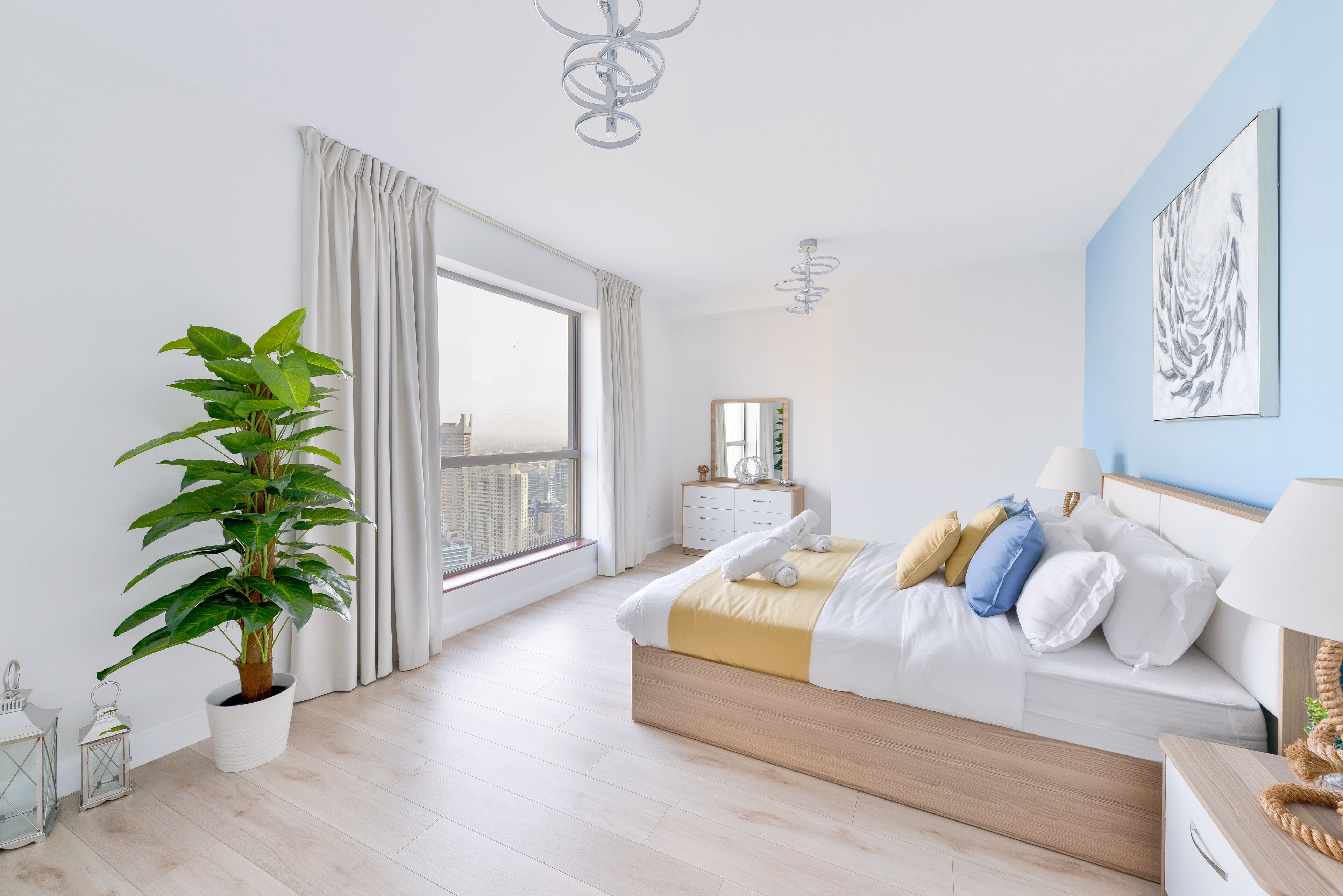 A modern bedroom with a large window, a potted plant, a bedside table, and a bed with white bedding and decorative pillows.