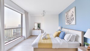 Un dormitorio moderno con un ventanal grande, cama con ropa de cama blanca y un detalle amarillo, una mesita de noche con lámpara y vista a la ciudad.