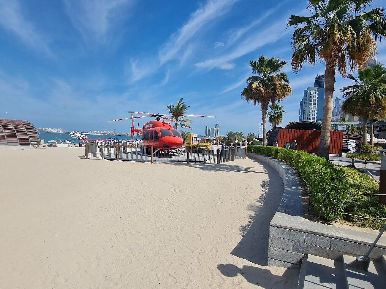 A red helicopter parked on a sandy beach with palm trees and a cityscape in the background.