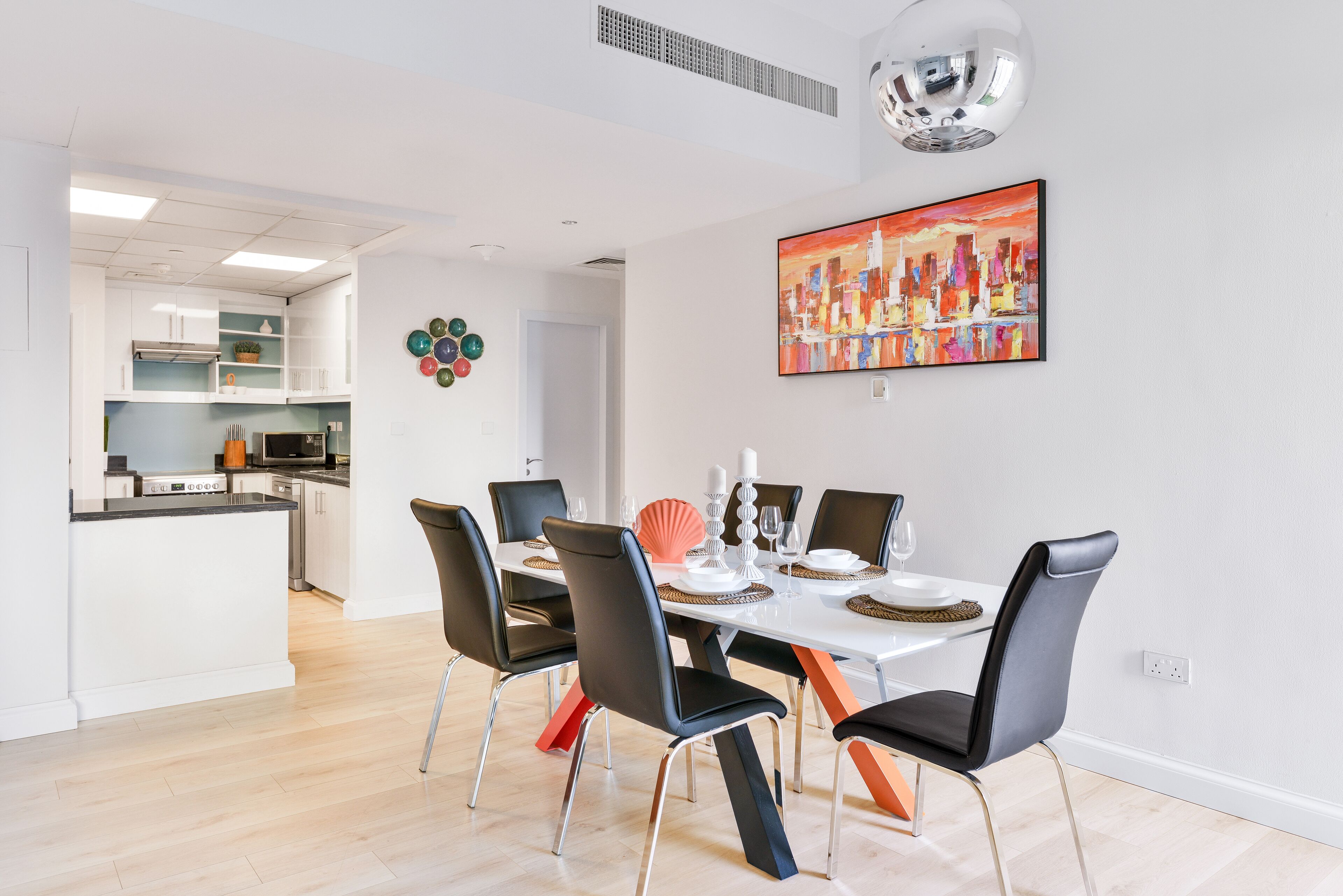 A modern dining area with a white table set for four, black chairs, and a cityscape painting on the wall.