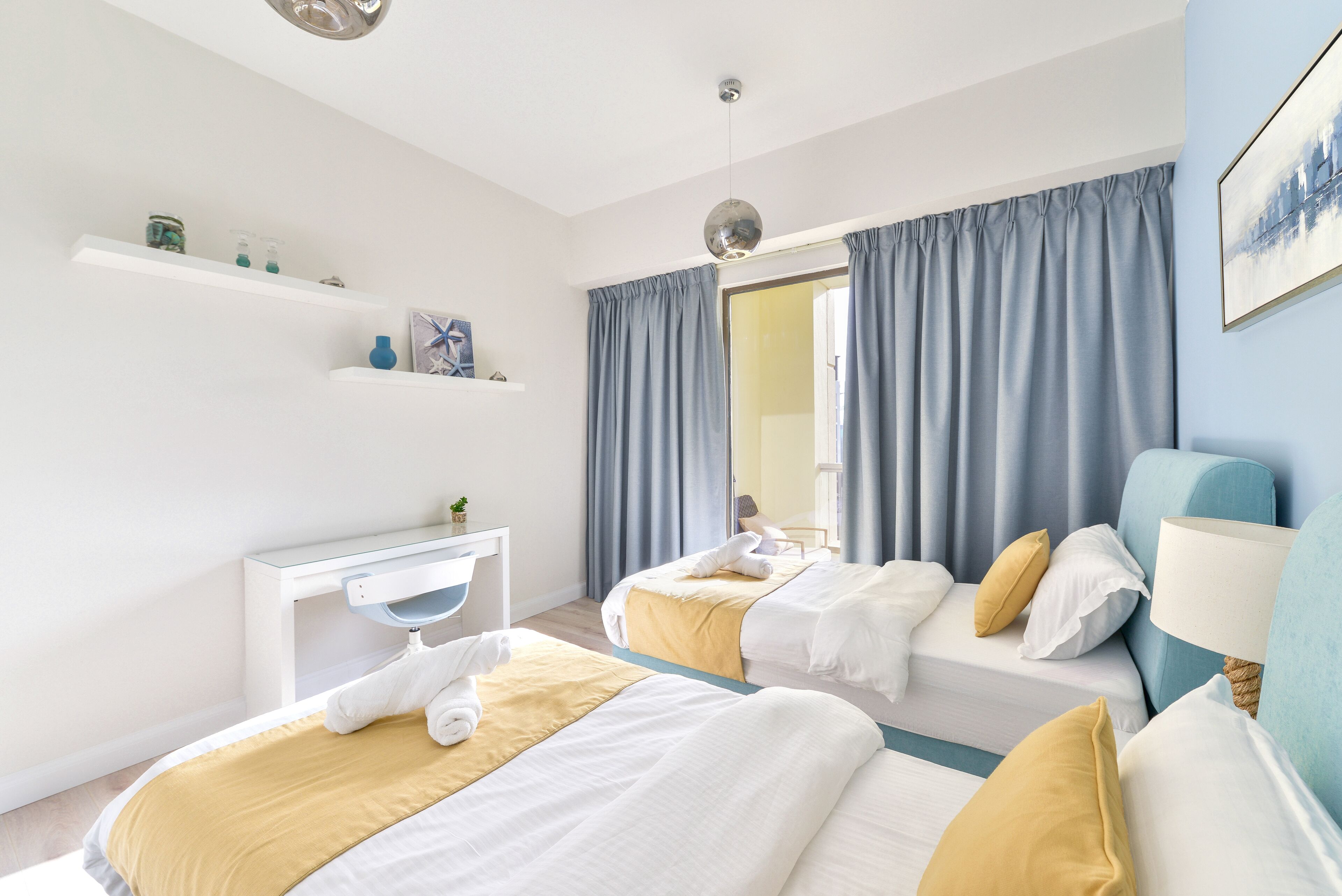 A hotel room with two beds, a desk, a chair, a lamp, and a window with curtains.