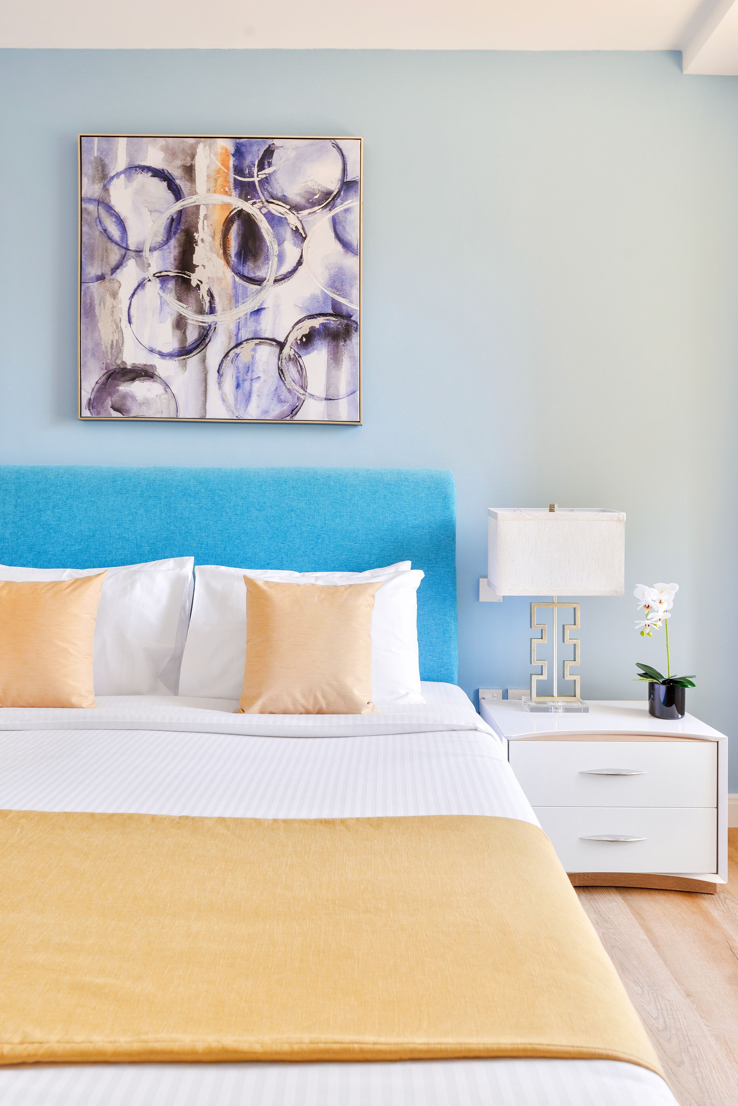 A neatly made bed with white and yellow bedding, a blue headboard, a bedside table with a lamp, and a wall-mounted abstract painting.