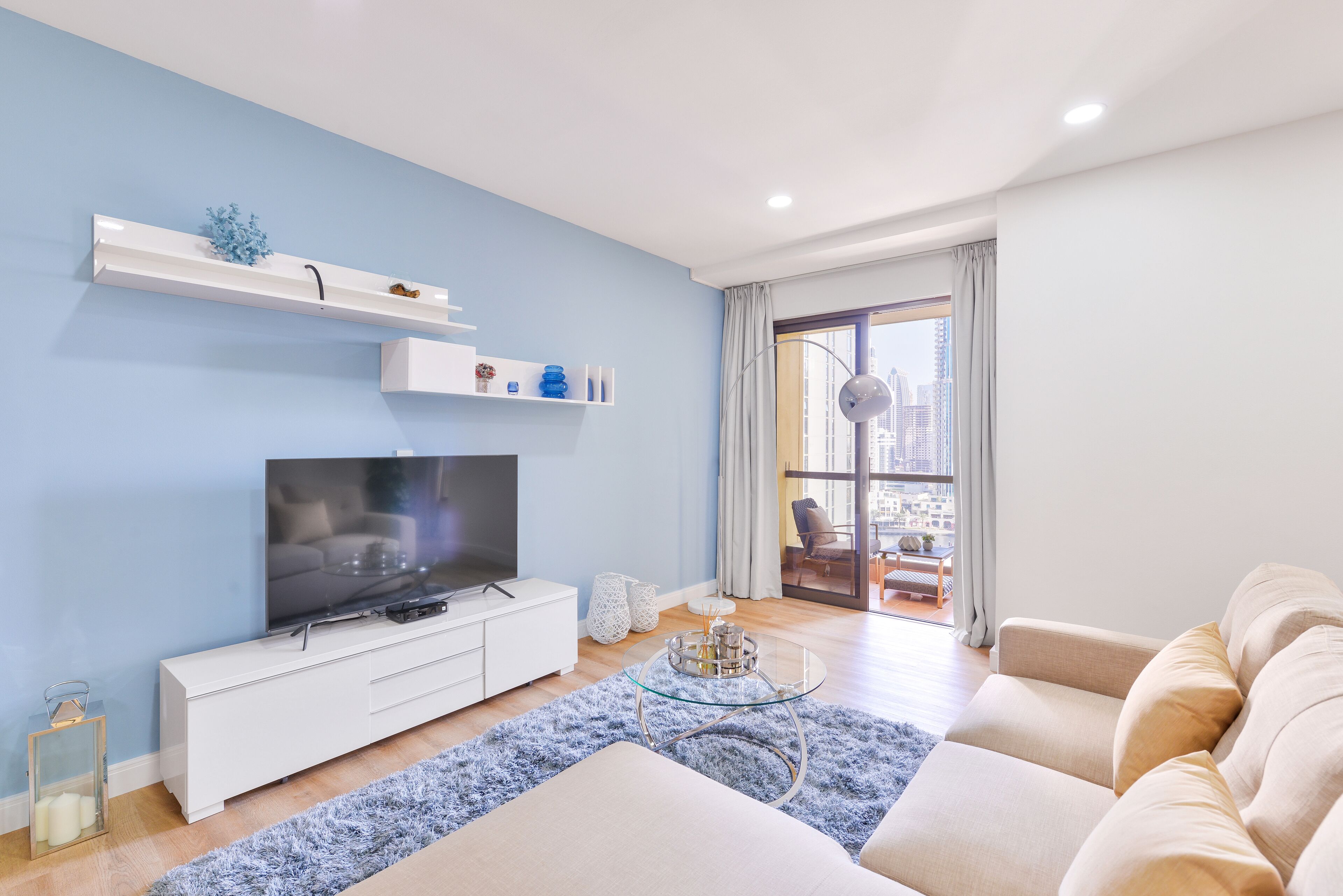 A modern living room with a flat-screen TV on a white cabinet, a glass coffee table, a beige sofa, and a city view through a large window.
