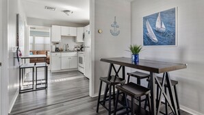 A modern kitchen with white cabinets, a dining area with a dark wooden table and stools, and a painting of sailboats on the wall. - Coastal Cottage~Fenced Yard~Fire Pit~Pet Friendly! (Villas)
