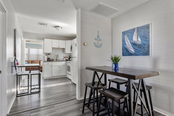 A modern kitchen with white cabinets, a dining area with a dark wooden table and stools, and a painting of sailboats on the wall.