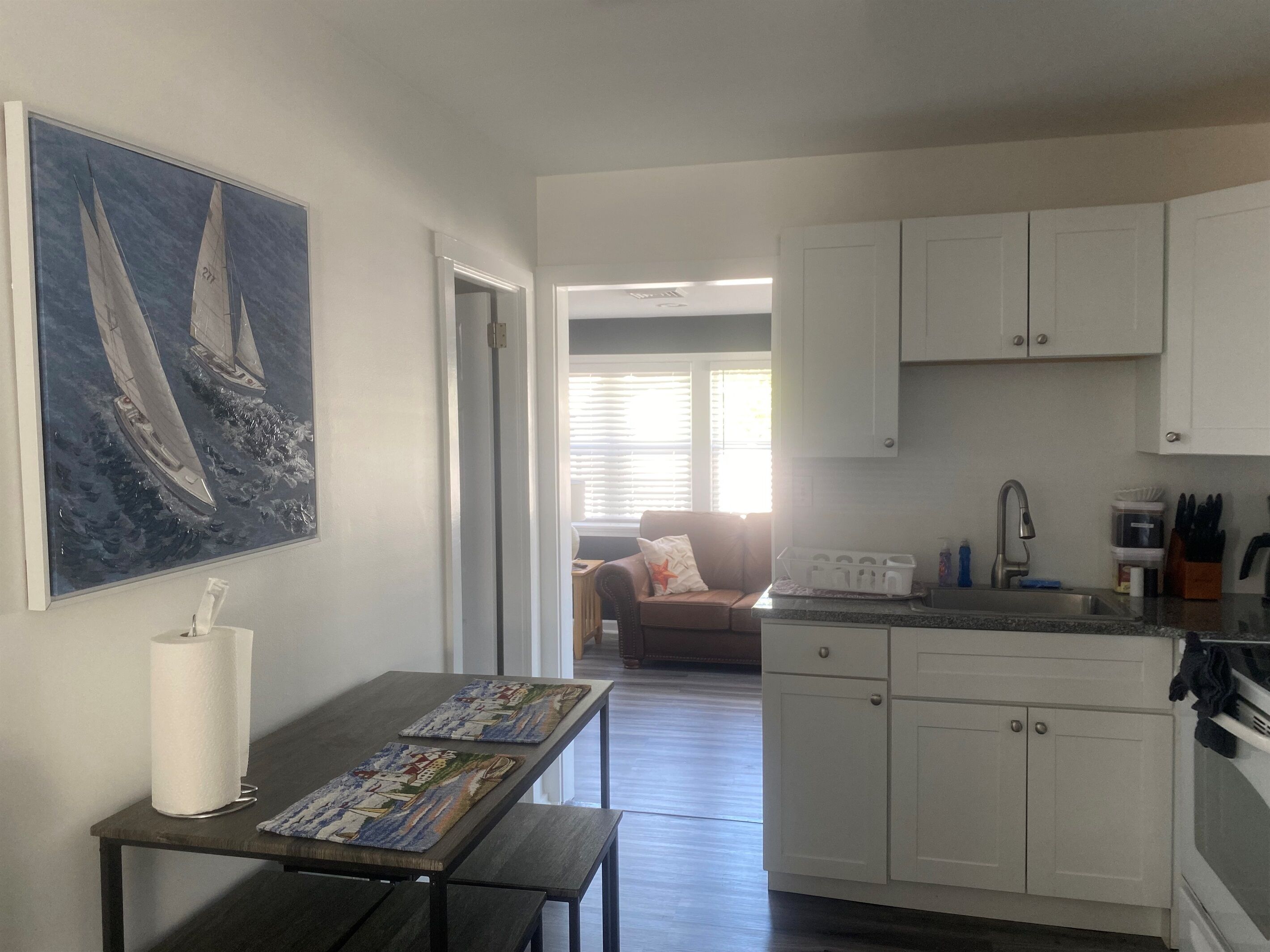 A compact kitchen with white cabinets, a dining table with placemats, and a painting of sailboats on the wall.