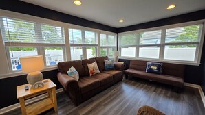 A living room with a brown leather sofa, a wicker chair, and a wooden table with a lamp. Large windows provide a view of the outside. - Coastal Cottage~Fenced Yard~Fire Pit~Pet Friendly! (Villas)