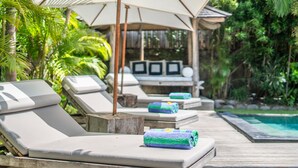 Une terrasse en bois avec des chaises longues et des parasols, une piscine et une végétation luxuriante.