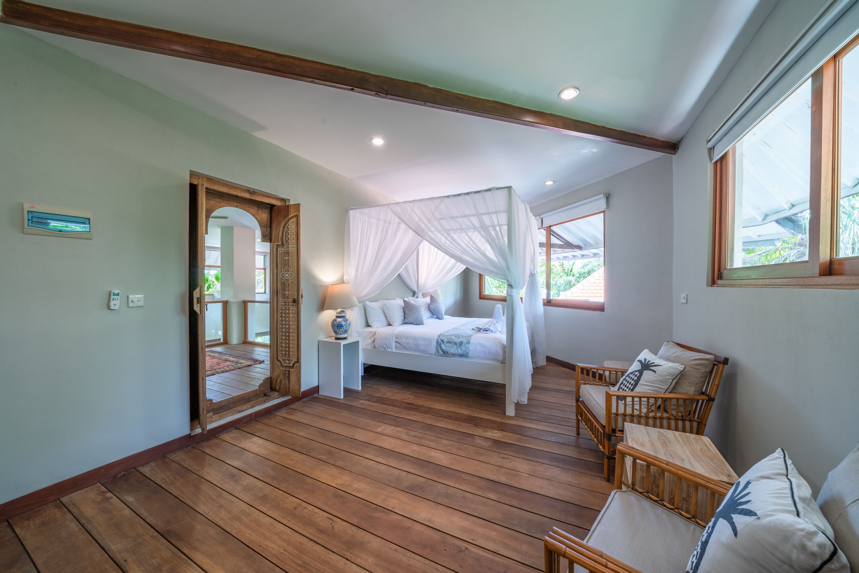 A bedroom with a four-poster bed, wooden flooring, and a view of greenery outside.