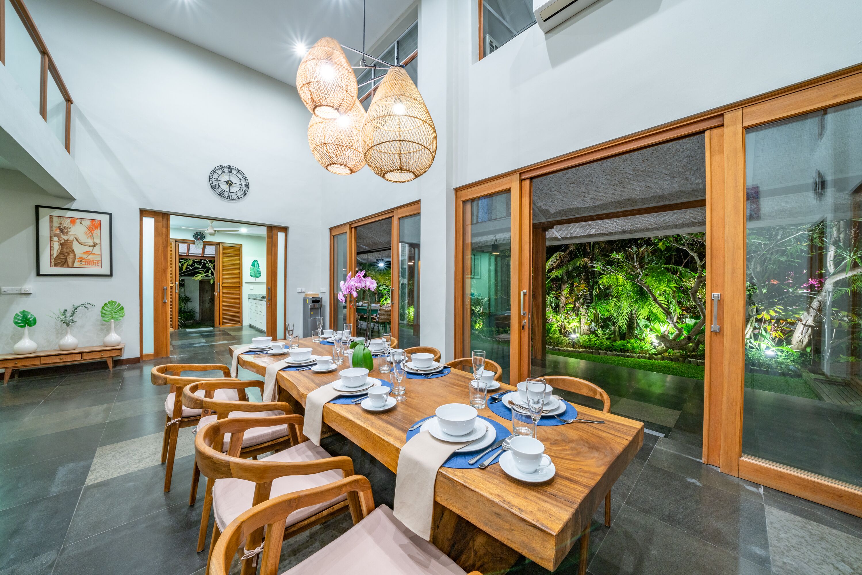 A dining area with a long wooden table set for six, surrounded by wooden chairs. There are large windows with a view of a lush garden.