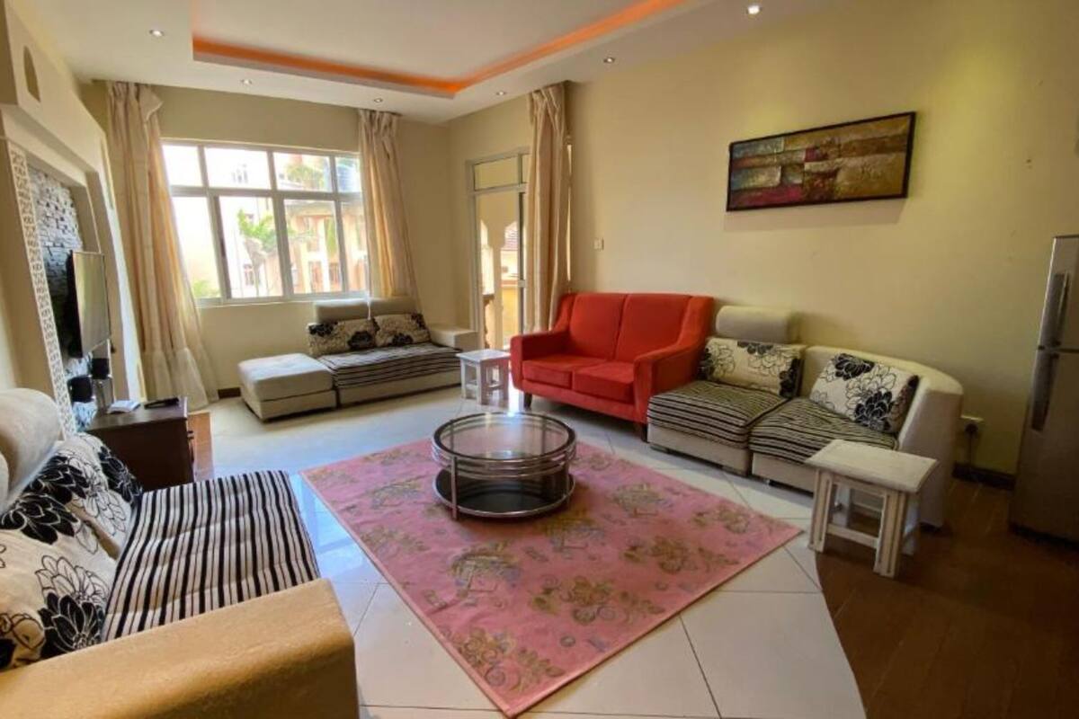 A living room with a sofa, armchair, ottoman, and a coffee table. There is a window with curtains, a painting on the wall, and a refrigerator in the corner.