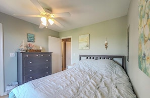 Une chambre à coucher comprenant un lit, une commode, un ventilateur de plafond, des œuvres d’art et une porte donnant sur une autre pièce.