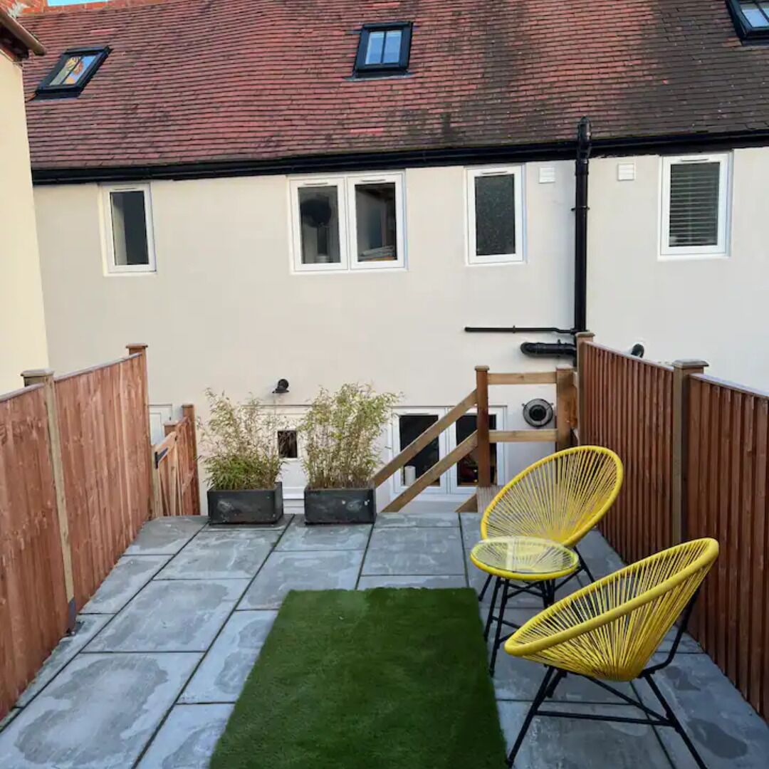 A patio with a green artificial turf, two yellow chairs, and a wooden staircase leading to a building with multiple windows and a red roof.