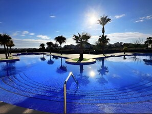 A large, clear blue swimming pool with a central island and palm trees, surrounded by a well-maintained landscape. - 2 Bed 2 Bath Penthouse on PGA Golf Course  (Sucina)