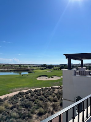 Uitzicht op een golfbaan met een vijver, een wit gebouw met balkon en een heldere blauwe lucht.