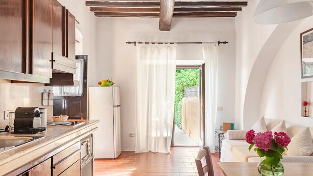 A kitchen with wooden cabinets, a white refrigerator, and a coffee machine. There is a dining area with a table and chairs, and a vase with flowers on the table.