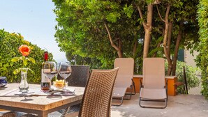 An outdoor dining area with wicker chairs and a table set for two, surrounded by lush greenery.