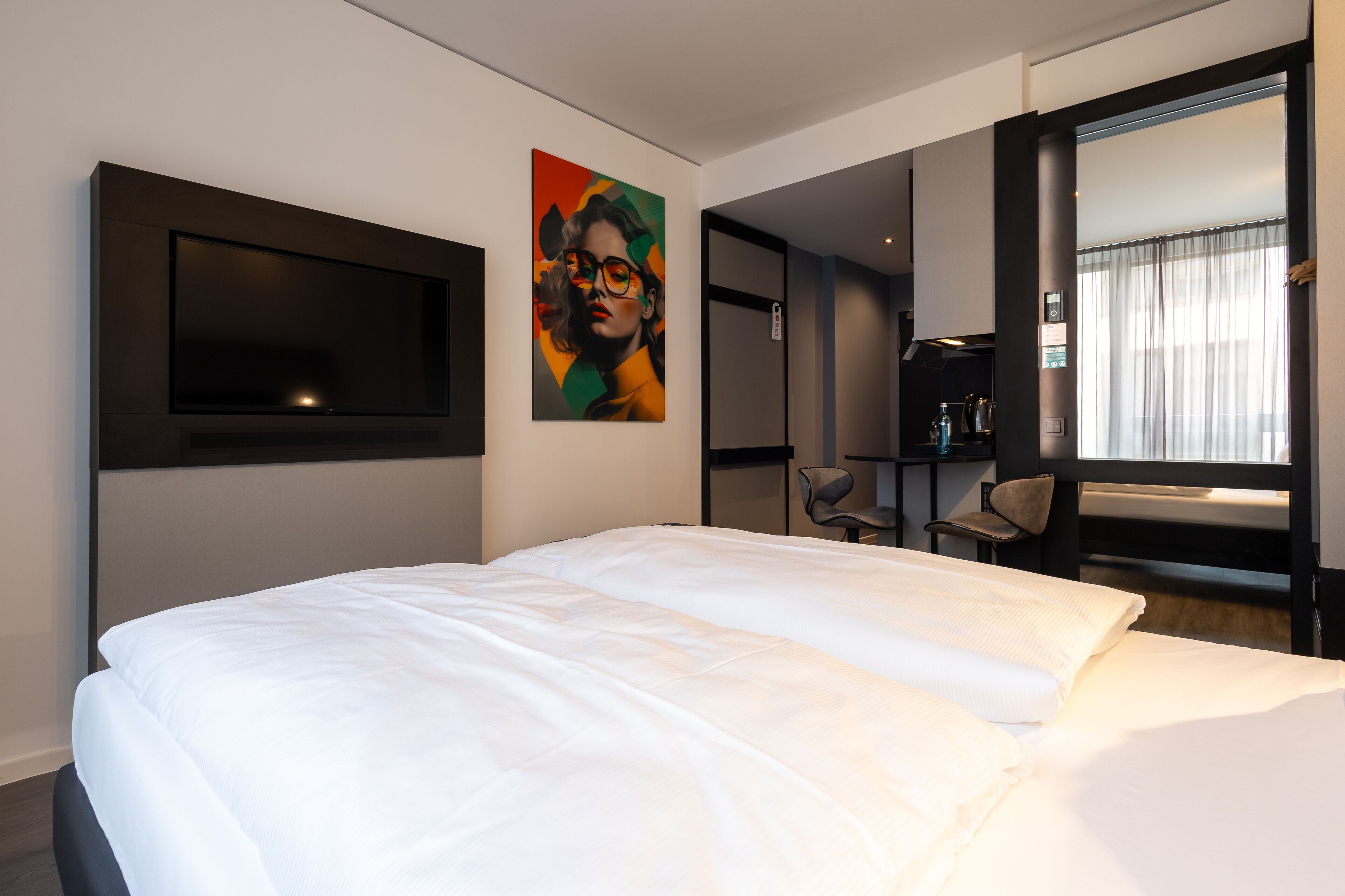 Ein modernes Hotelzimmer mit einem großen Bett, einem flachbildfernseher an der Wand, einem farbenfrohen Gemälde und einer Sitzecke mit Stühlen und einem Tisch.