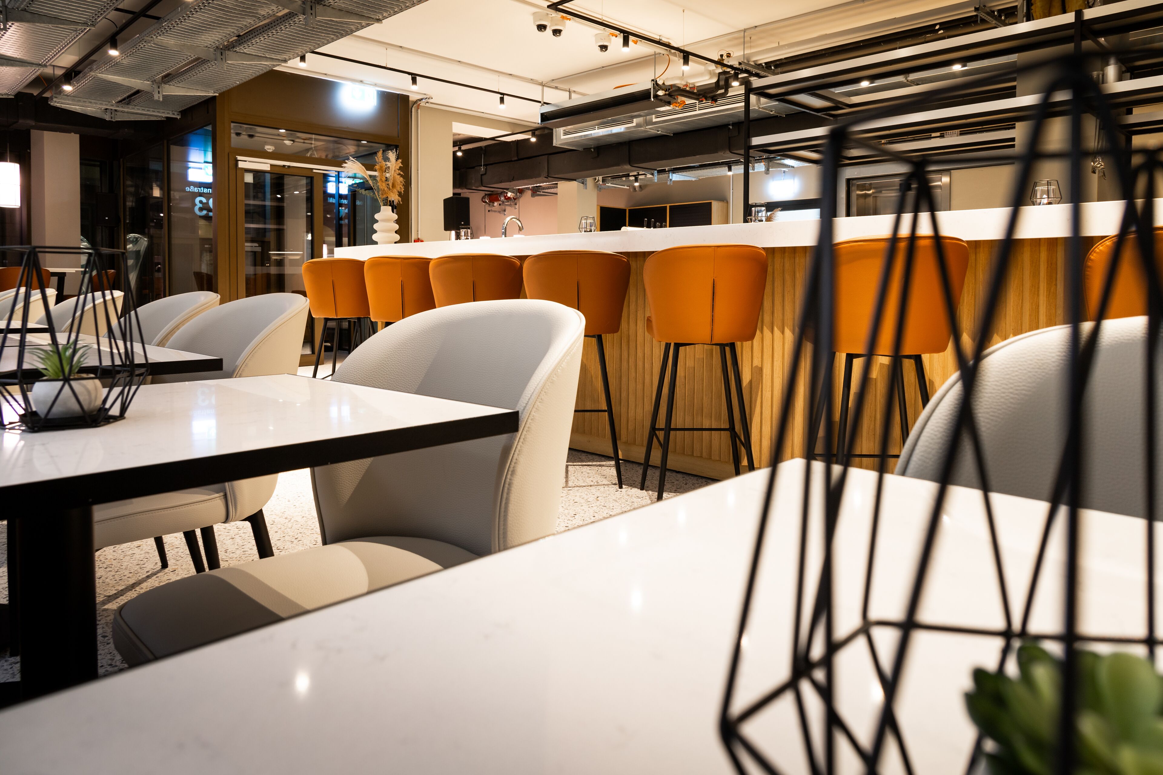 A modern dining area with white tables and orange chairs, a bar with stools, and a kitchen in the background.
