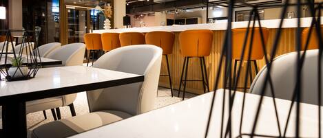 A modern dining area with white tables and orange chairs, a bar with stools, and a kitchen in the background.