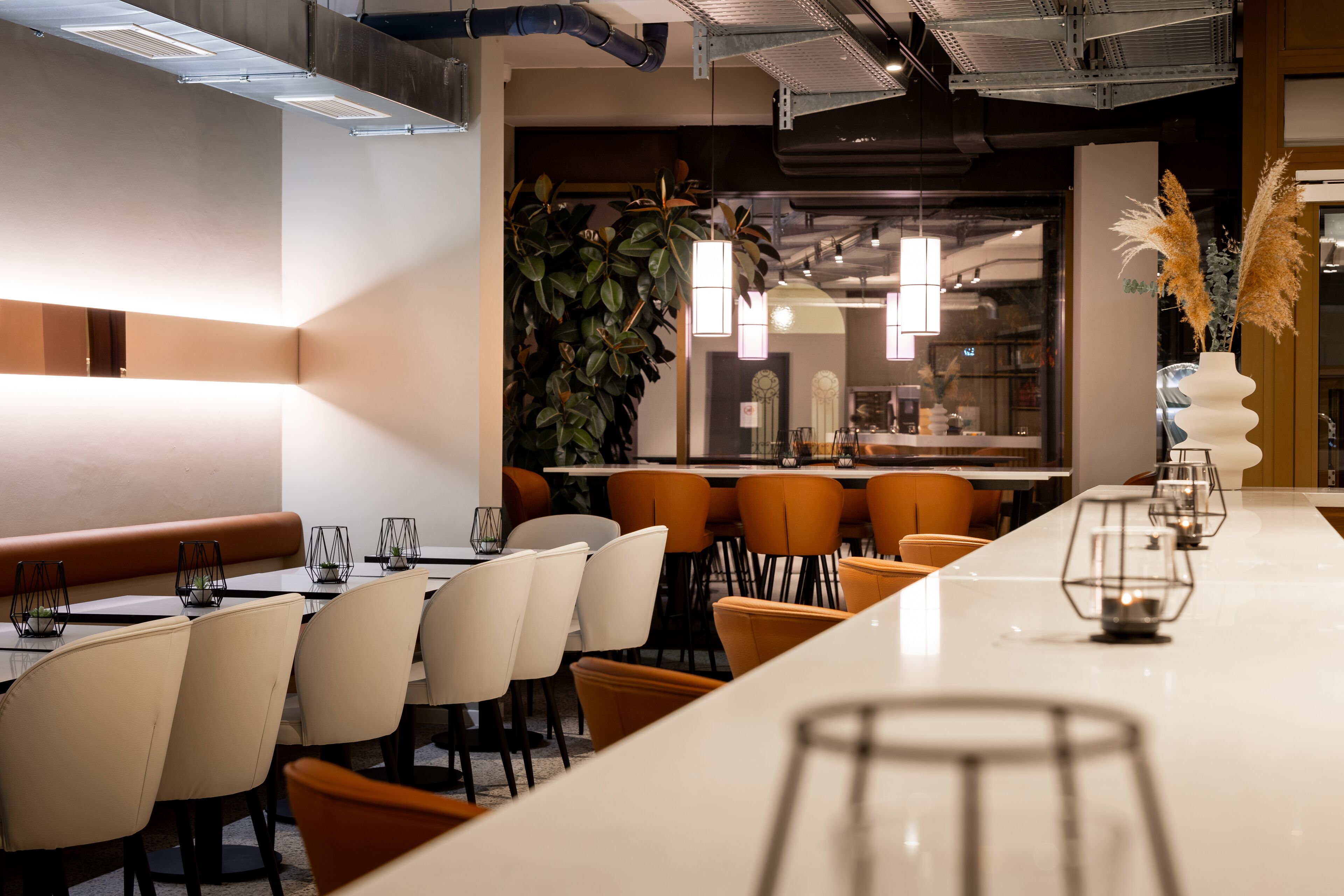 A modern restaurant with a long white counter, multiple orange chairs, and a large green plant in the background.
