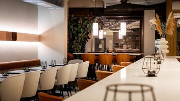 A modern restaurant with a long white counter, multiple orange chairs, and a large green plant in the background.