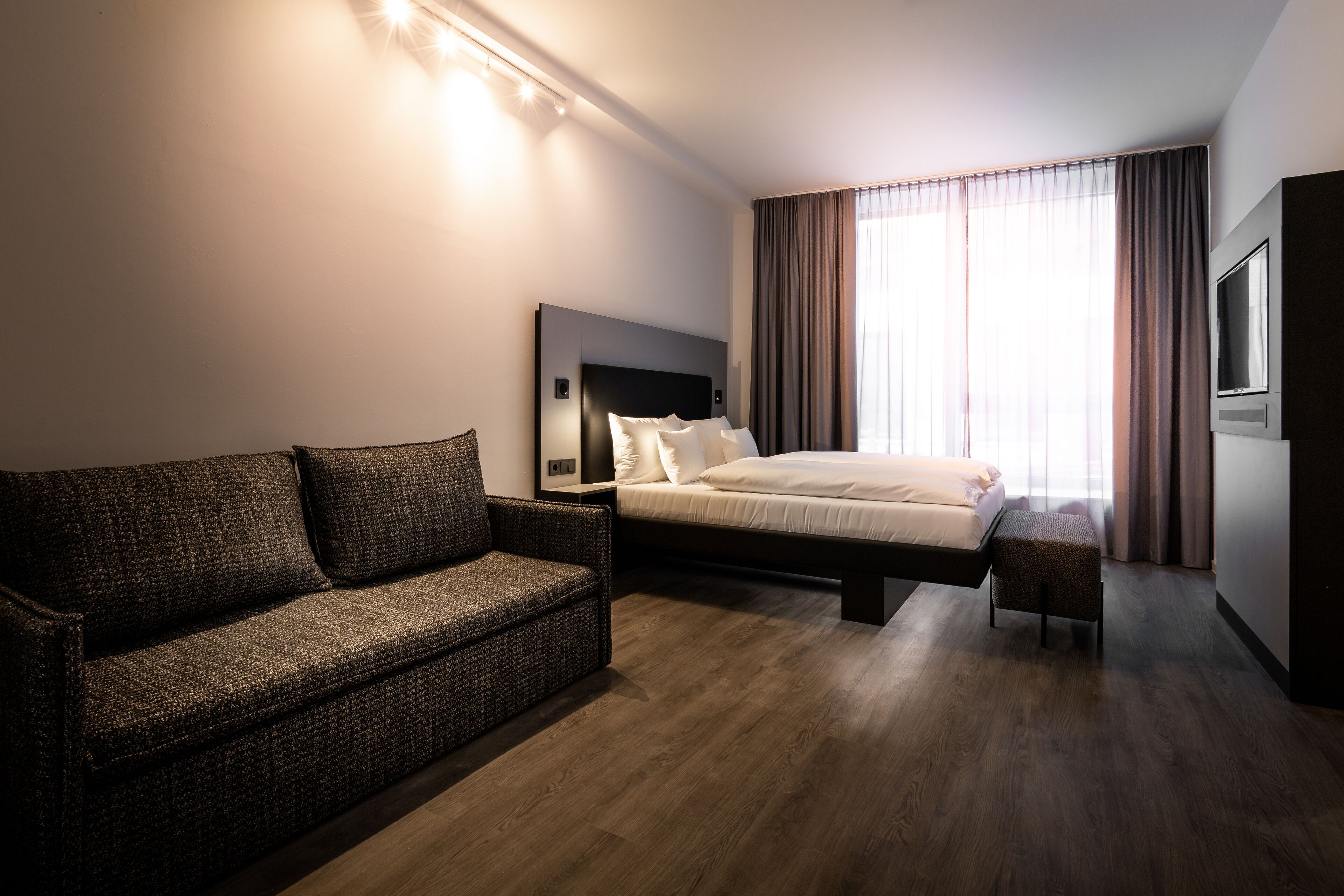 Une chambre d’hôtel moderne avec un grand lit, un canapé, une télévision et une grande fenêtre avec des rideaux.
