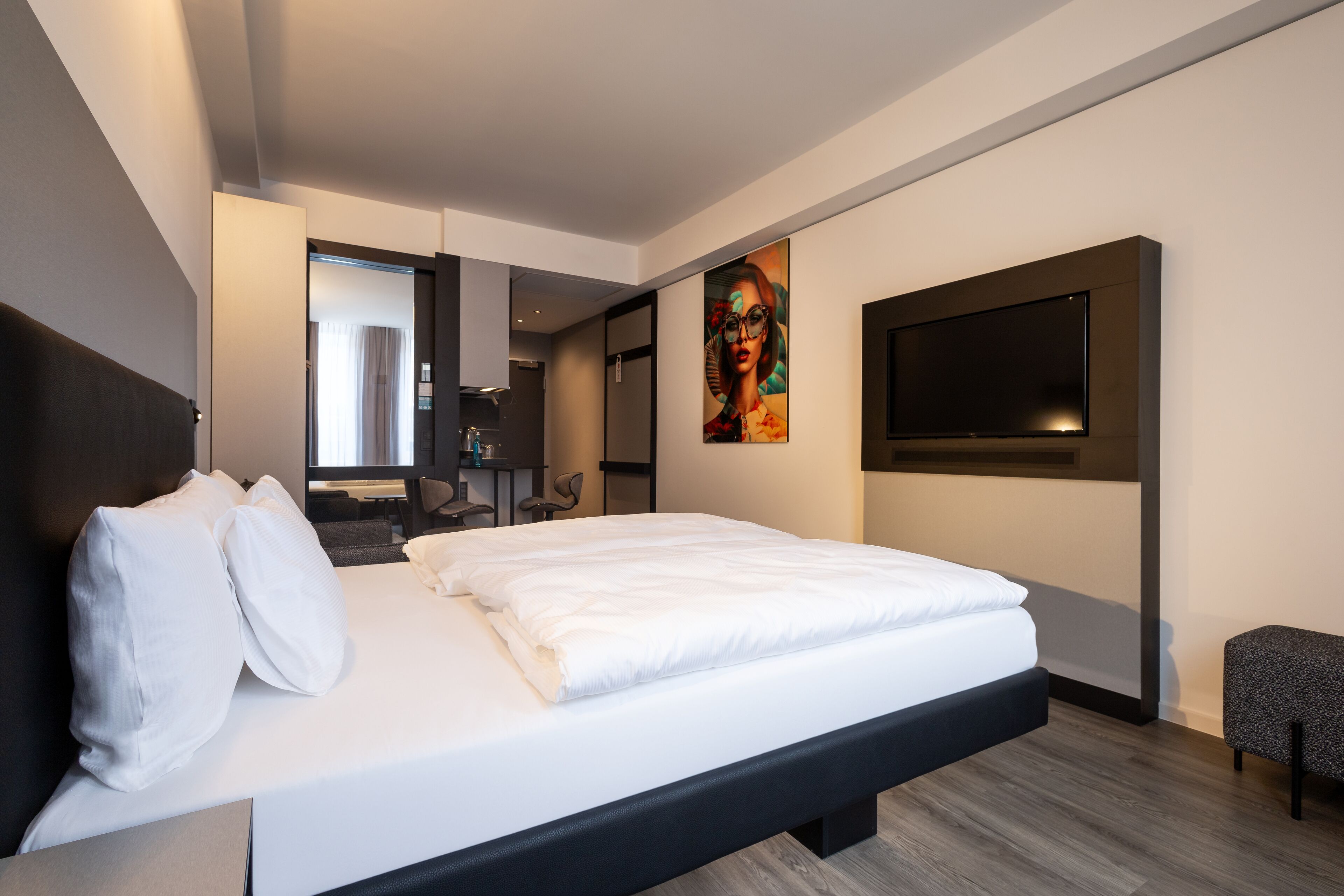 A modern hotel room with a large bed, a flat-screen TV mounted on the wall, a painting above the bed, and a small seating area.