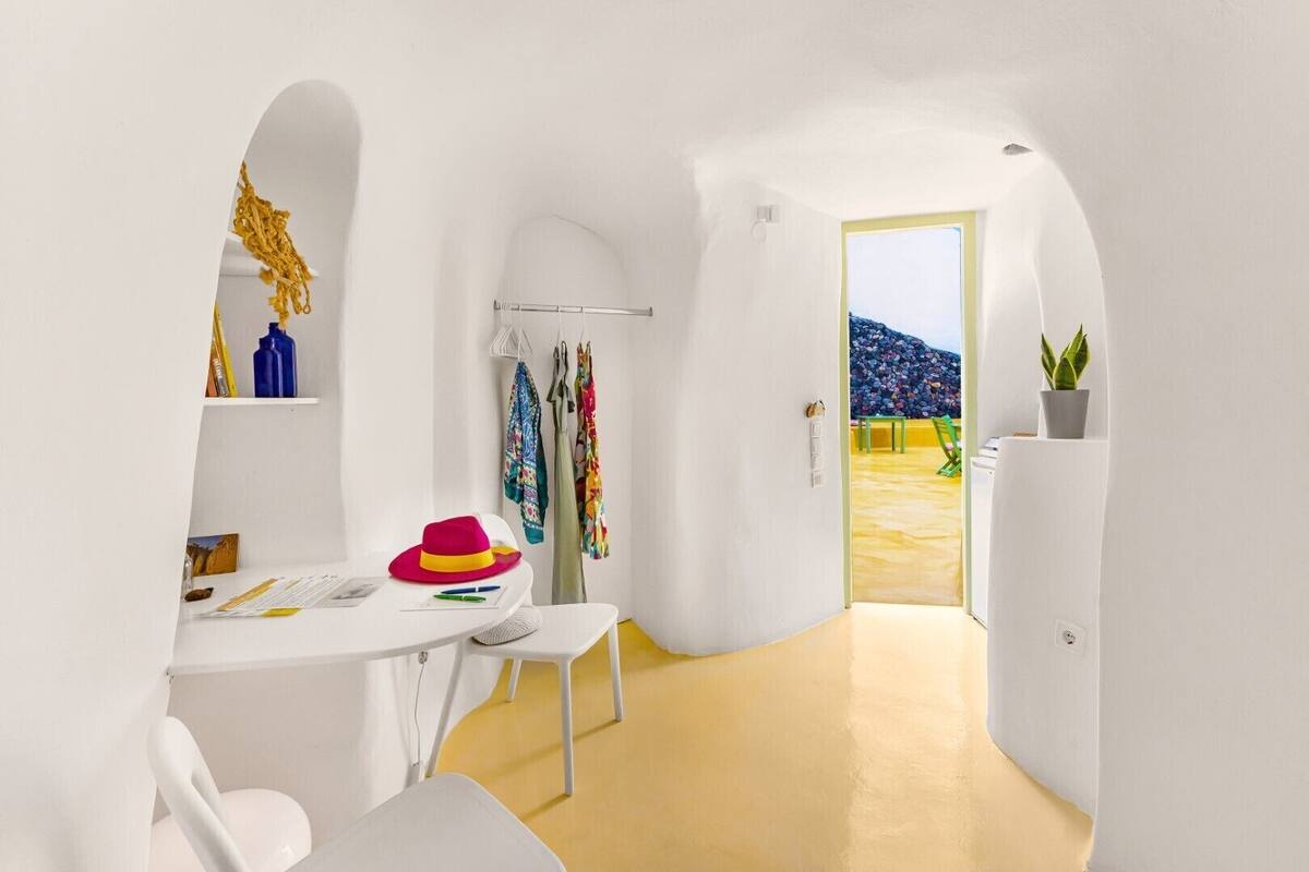 A modern, white-walled room with a yellow floor, a small table with a hat, and a door leading to an outdoor area with a yellow wall.