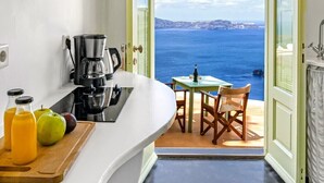 A kitchen with a coffee maker, a cutting board with fruits, and a view of the sea through an open door. - Sensyo Caves (Santorini)