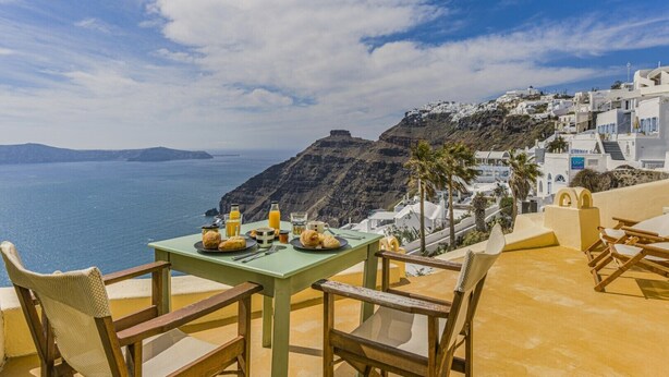 A table set with breakfast items overlooking a coastal view with white buildings and a cliff. - Sensyo Caves (Santorini)