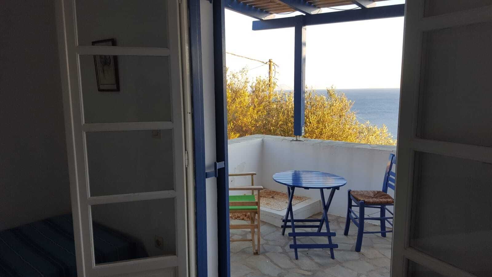 Une porte ouverte donnant sur un balcon équipé d’une table et de chaises, d’où l’on peut admirer un plan d’eau et des arbres.