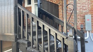 Een houten trap met metalen leuning die naar een gebouw van baksteen leidt, met ramen en een deur.