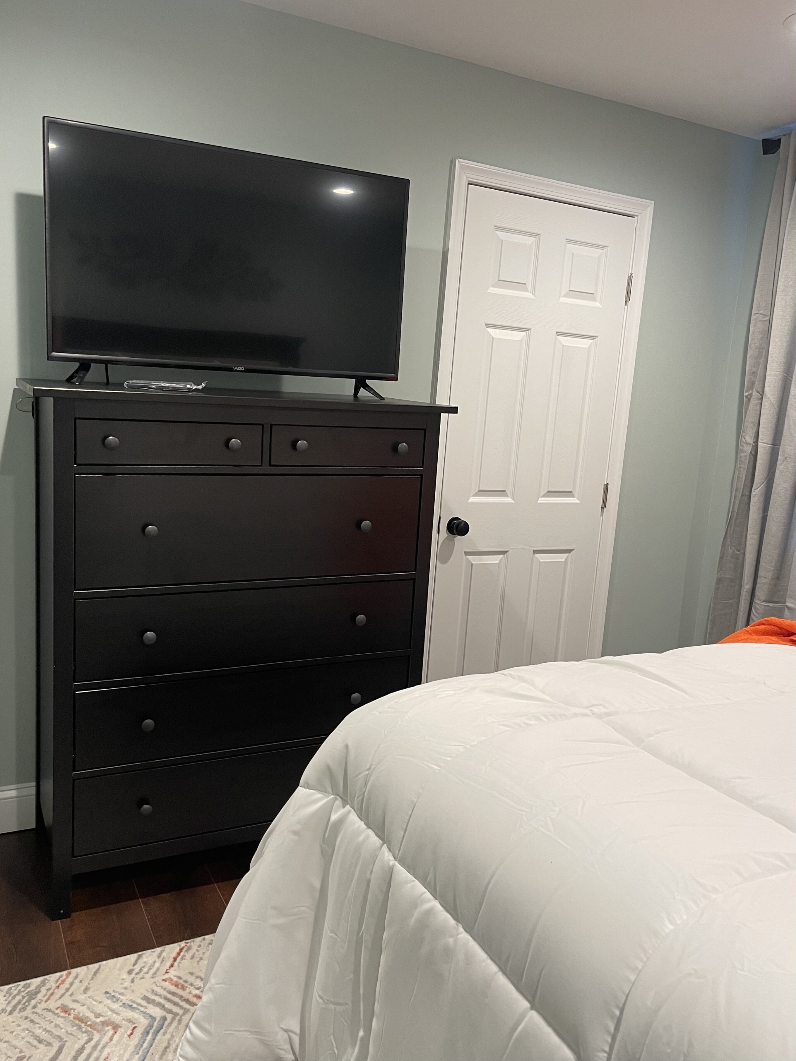 A bedroom with a black dresser, a television on top, a white door, and a bed with white bedding.