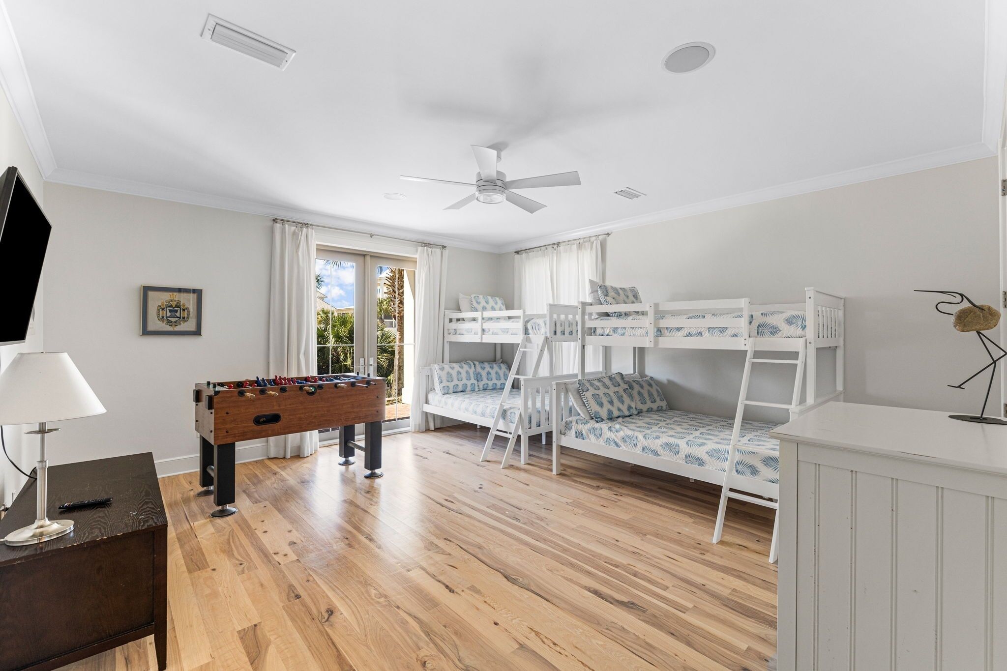 A room with a wooden floor, a foosball table, bunk beds with patterned bedding, a ceiling fan, and a window with curtains.