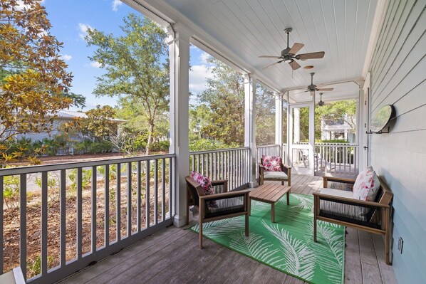 A covered porch with wooden furniture, a green rug, and a view of trees and a house.