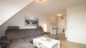 A modern living room with a grey sectional sofa, a white coffee table with a plant, and a painting of a coastal scene on the wall. - TOP vacation apartment for up to 2 people. Bright and modern furnishings. (Hörnum)
