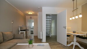 A modern living room with a sofa, a coffee table, and a dining area with a table and chairs. A staircase is visible in the background leading to another floor. - TOP vacation apartment for up to 2 people. Bright and modern furnishings. (Hörnum)